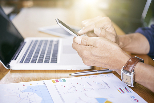 Close-up of businessman sitting at the table with laptop and documents and communicating online using his smartphone