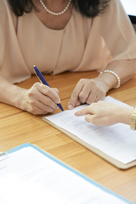 Close-up of mature businesswoman sitting at the table and signing a business contract with pen with her partner pointing at document
