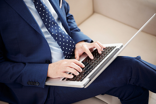 Close-up of businessman in suit sitting on sofa and typing on keyboard of laptop computer at office
