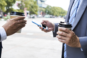 Close-up of two businessmen in suit standing in the city drinking coffee from disposable cups and using smartphone