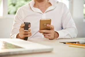 Close-up of young businessman sitting at the table with wristwatch and mobile phone and checking time on both gadgets at office