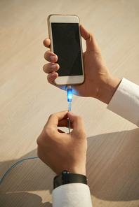 Close-up of young man connecting cable to mobile phone to charge it at office
