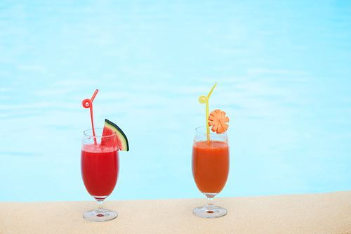 Still life of two glasses of exotic cocktails standing on edge of outdoor swimming pool,  blue water surface on background
