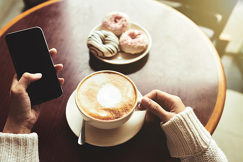 Hands of woman having cup of cappuccino with doughnuts and checking social media on smartphone