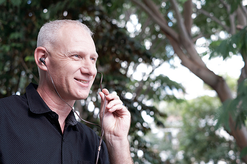 Good-looking mature Caucasian man smiling cheerfully while sitting outdoors with earphones and talking on mobile phone