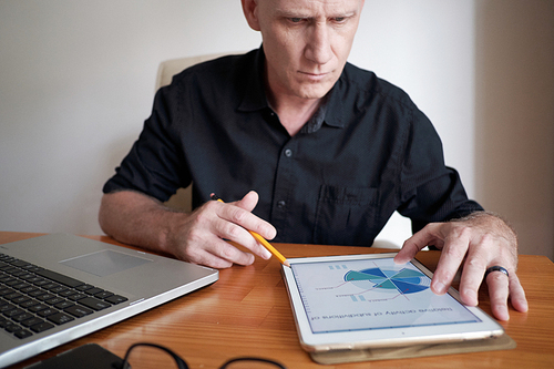 Concentrated senior Caucasian man sitting at office desk and looking at digital tablet with chart while analyzing market situation