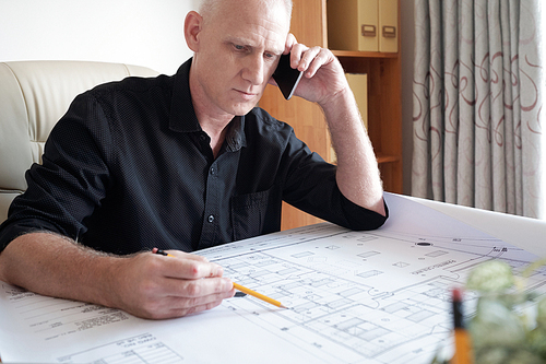 Concentrated middle-aged Caucasian man sitting at office table, examining blueprint of house and consulting with colleague on smartphone