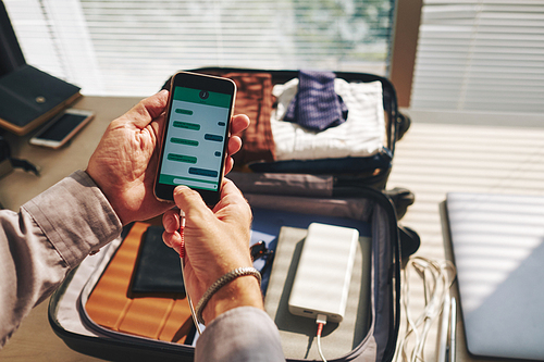 Hands of man texting friend when packing suitcase for vacation or business trip