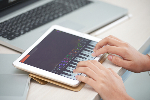 Crop view of elegant hands of woman sitting at desk with laptop and learning to play piano using tablet app