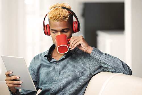 Handsome young man in headphones drinking coffee and reading data on tablet computer