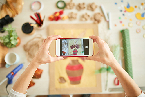 Close-up shot of creative woman taking picture of handmade decor item with help of smartphone while sitting at desk