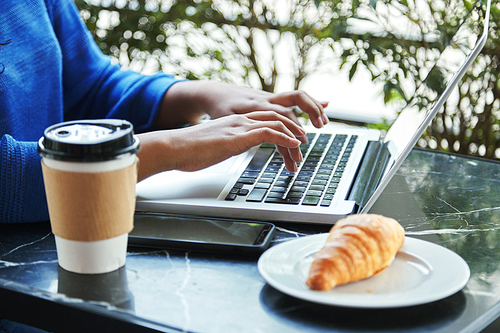 Crop Indonesian woman sitting at cafe table and typing on laptop keyboard near fresh croissant and cup of hot beverage