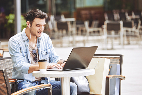 Cheerful young man sitting at table in outdoor cafe and working on laptop