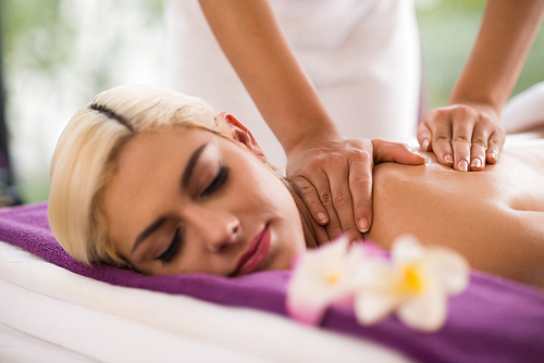 Relaxed blond-haired woman with eyes closed lying on massage table and enjoying procedure, blurred background