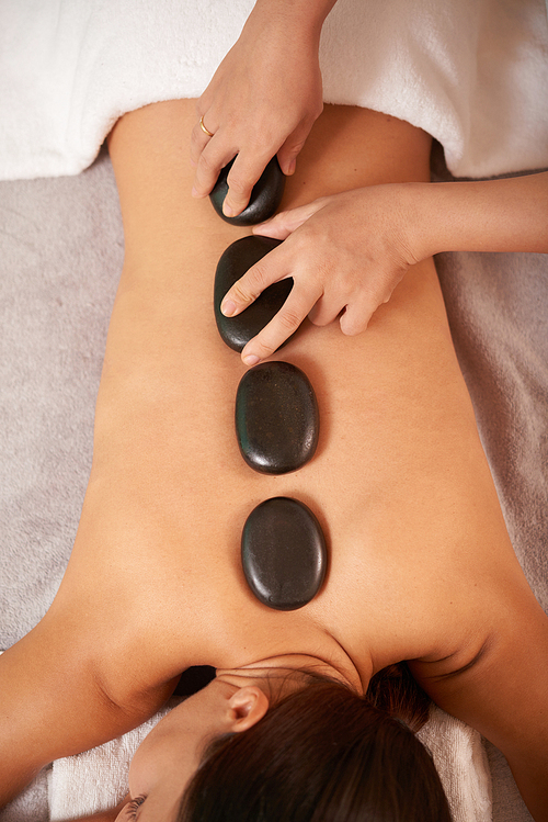 Overview of relaxed client back and hands of cosmetician putting spa stones before procedure
