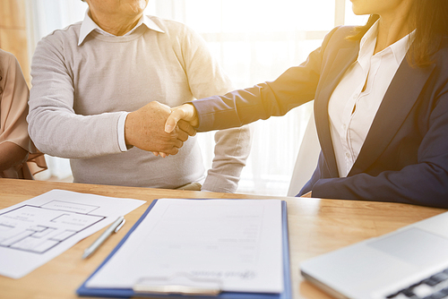 Close-up shot of unrecognizable senior man and estate agent shaking hands after successful completion of meeting, purchase agreement lying on table