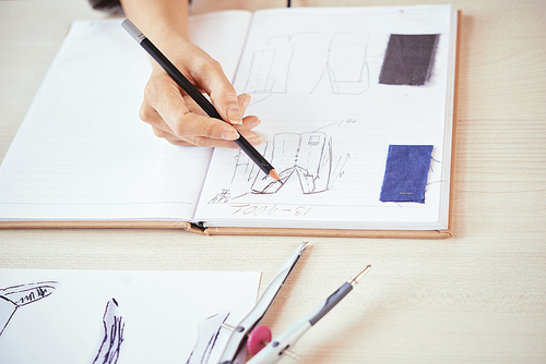 Crop hand of fashion designer drawing sketches of stylish clothes near samples of fabric in open notebook