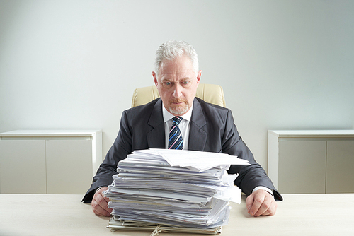 Shocked senior manager wearing classical suit looking at pile of documents while sitting at office desk, waist-up portrait shot