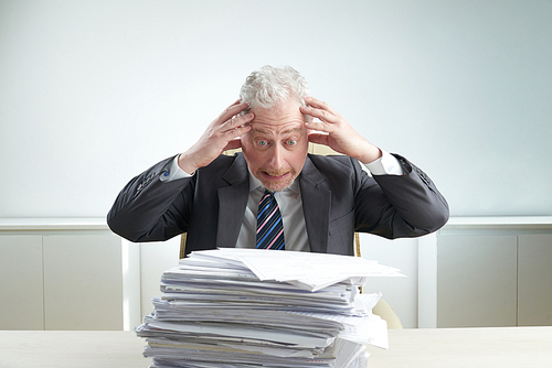 Stressed senior businessman wearing suit holding head and looking at pile of documents with anger while sitting at office desk, waist-up portrait shot