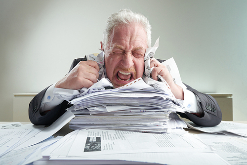 Portrait of mad senior entrepreneur resting his chin on pile of documents, screaming and crumpling lists of paper while relieving stress, office interior on background