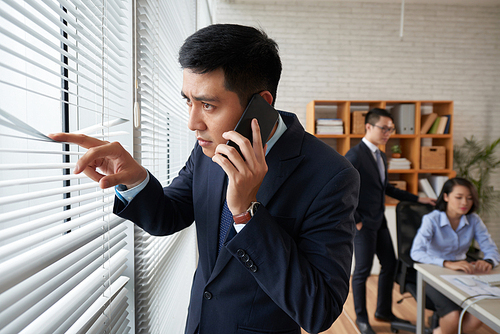 Young Vietnamese businessman looking through blinds on office window when calling on the phone