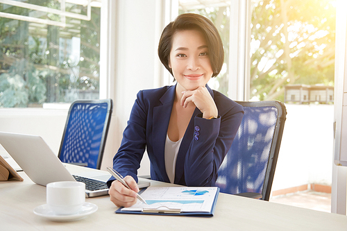 Portrait of smiling businesswoman sitting at the table with laptop and cup of tea and preparing financial report for presentation