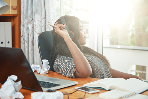 Side view of modern black woman in headphones sitting at table in office looking away in sunlight
