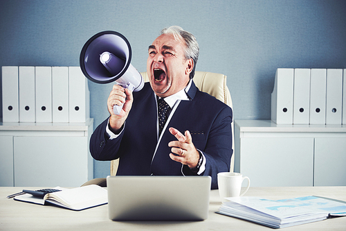 Aged male in black elegant business suit sitting at office desk with gadgets and documents and shouting fiercely into large megaphone looking up and pointing finger at camera.