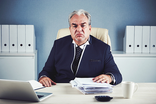 Bored sad mature male in dark elegant suit sitting at office workplace holding hand on large pile of documents and 