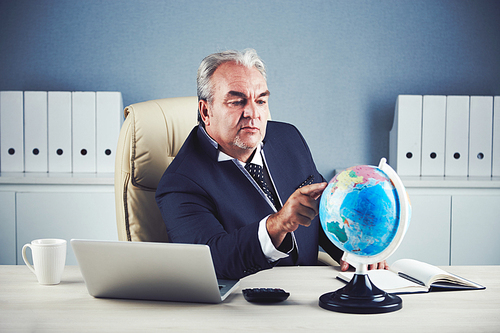 Senior male in dark business suit sitting comfortably at office desk and looking at world globe choosing location of future vacation