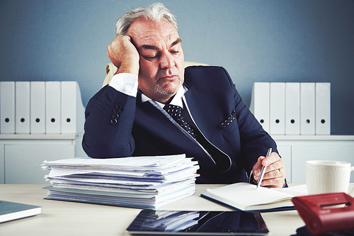 Bored senior male in dark elegant suit sitting at office workplace leaning on arm against large pile of documents and reading and writing in notepad on desk.