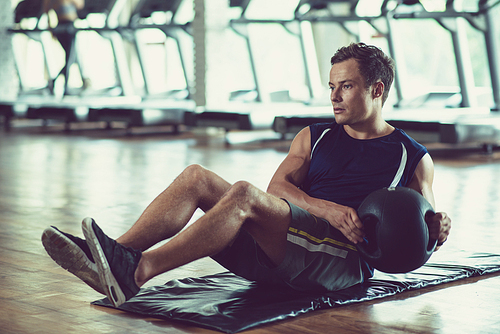 Confident young athlete sitting on mat and doing sit-ups with double handle medicine ball, interior of spacious gym on background