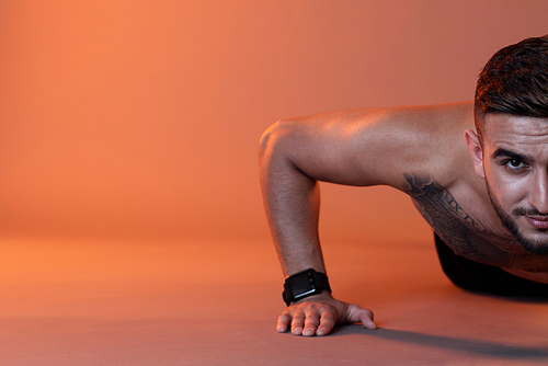 Crop tattooed shirtless man  while doing push-ups on floor against red illuminated background