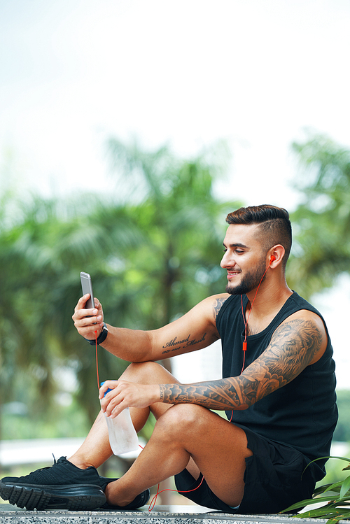 Handsome cheerful man in headphones sitting on ground outdoors and browsing smartphone in leisure having bottle of water