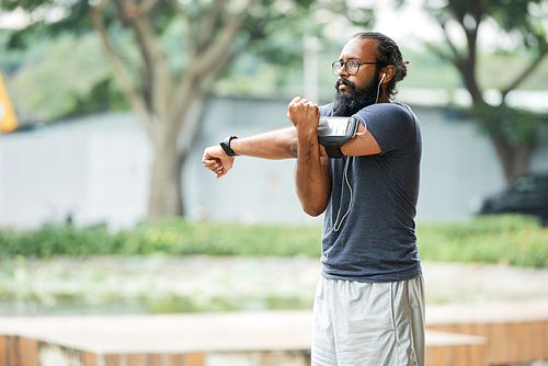 Athletic bearded Indian man in glasses doing physical exercise outdoors and listening to music through earbuds on blurred background