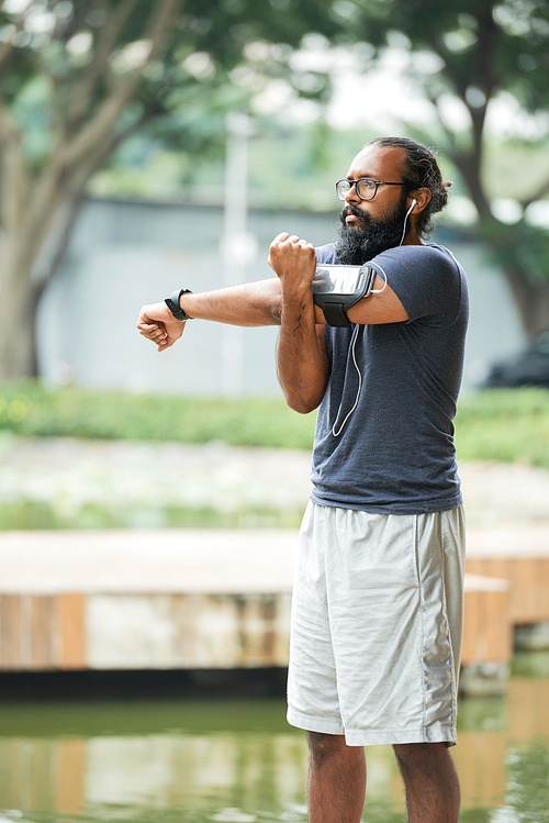 Bearded Indian sportsman in glasses and activewear listening to music with earphones and warming up outdoors on blurred background