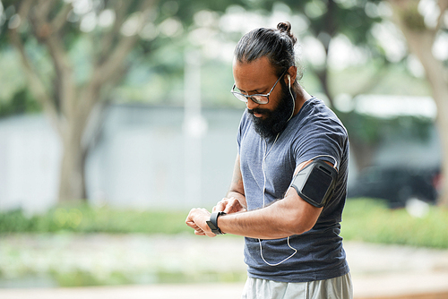 Bearded athletic Indian man in glasses checking time on smartwatch while having workout outdoors on blurred background