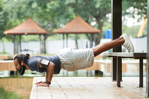 Bearded Indian sportsman with smartphone on his shoulder doing push-ups outdoors and listening to music with earphones