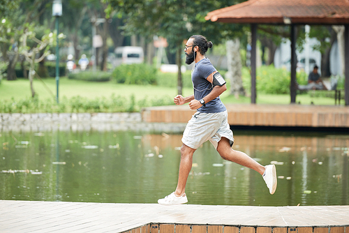 Side view of athletic bearded Indian man with smartphone on shoulder jogging outdoors and listening to music through earbuds