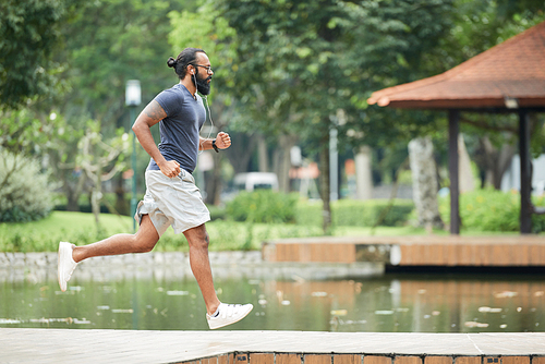 Side view of athletic bearded tattooed Indian man jogging in the morning outdoors and listening to music with earphones