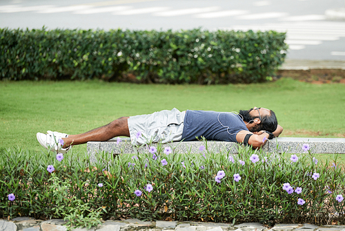 Athletic bearded Indian man enjoying recreation outdoors, lying on bench and listening to music through earphones after training