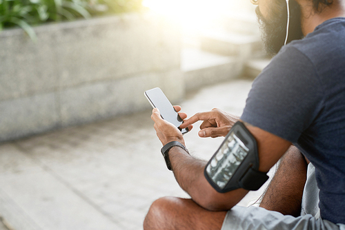 Close-up view of unrecognizable bearded man choosing song on smartphone while resting after training on sunny morning outdoors