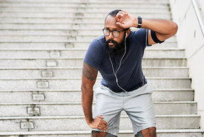 Bearded tattooed Indian male athlete standing on steps outdoors, wiping sweat from his forehead and recovering breath after running