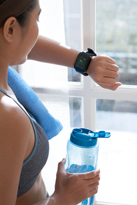 Side view of sportswoman holding bottle of water and checking smart watch in sunlight