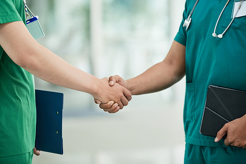 Two anonymous men in medical scrubs holding tablet and clipboard and shaking hands while standing on blurred background of hospital hall