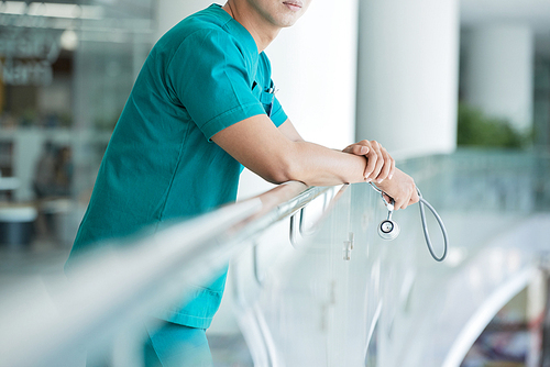 Unrecognizable health professional holding stethoscope and leaning on railing while standing in hospital hall