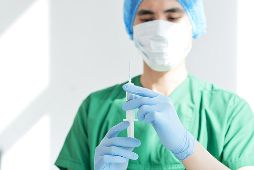 Asian health professional in mask holding syringe and preparing for injection while working in hospital
