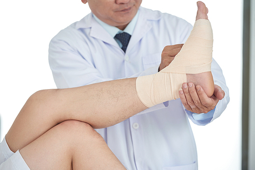 Unrecognizable medical practitioner fastening bandage on injured leg of crop man while working in office