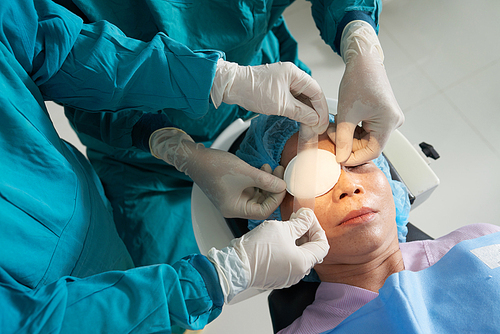 Crop shot from above of careful surgeons applying patch with tape on eye of mature Asian patient after surgery