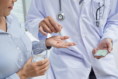 Close-up shot of female patient holding glass of water in hand while doctor wearing white coat giving her pills